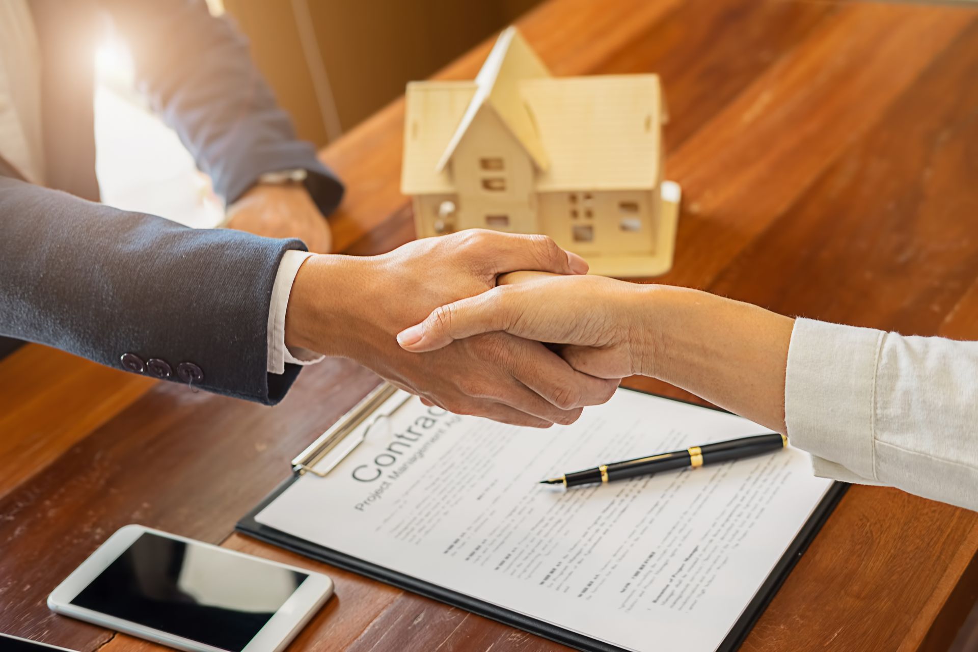 Handshake over a contract, a small house model in the background, signaling a real estate deal.