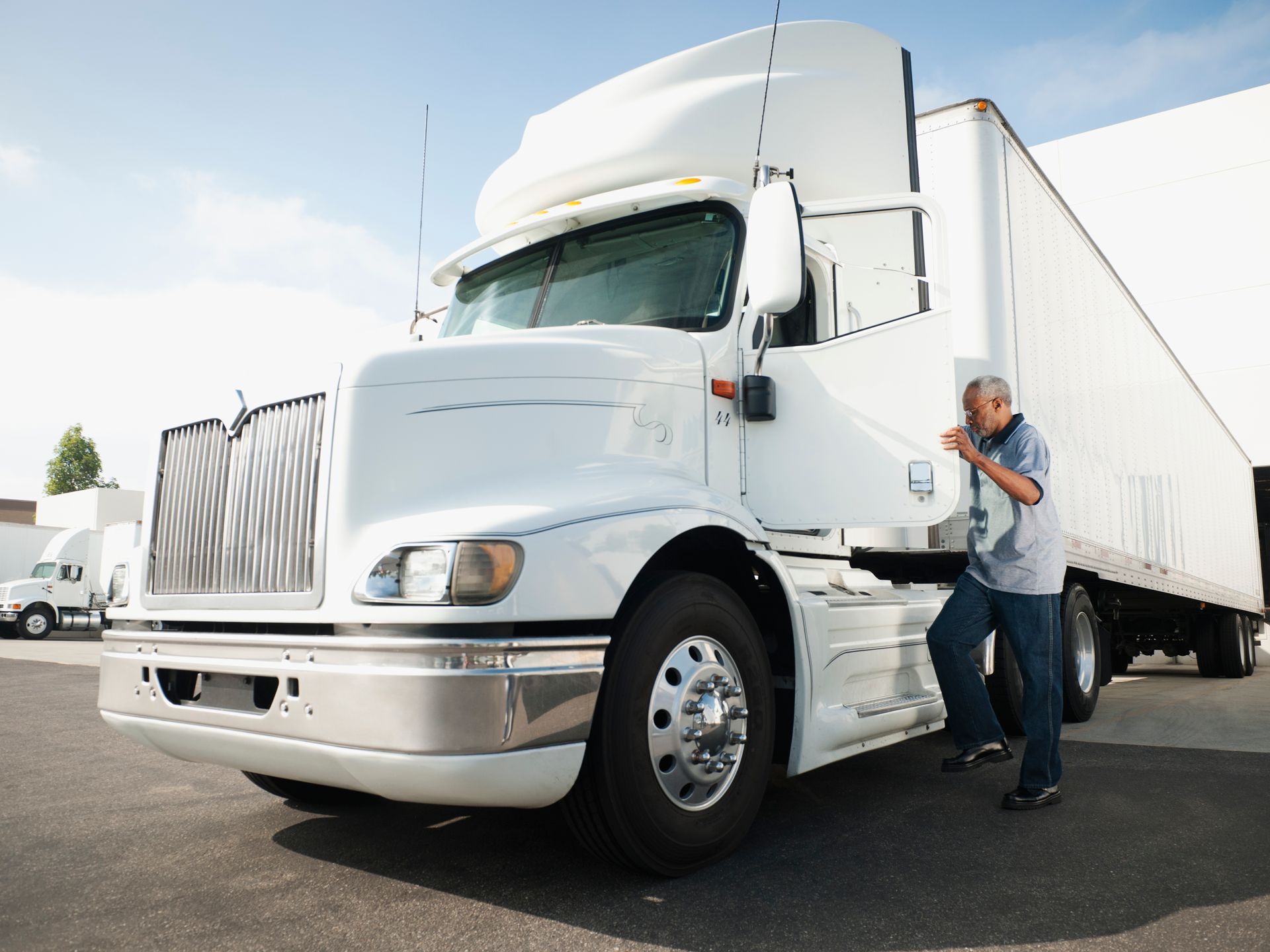 White semi-truck with a driver getting into the cab in a paved lot on a sunny day.