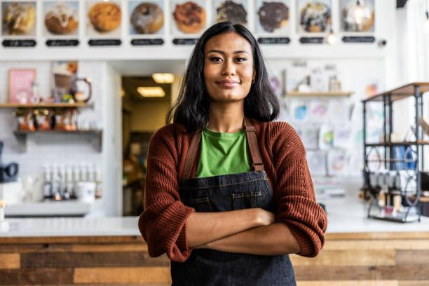 Woman with arms crossed, in a coffee shop, wearing an apron and brown sweater, smiling.