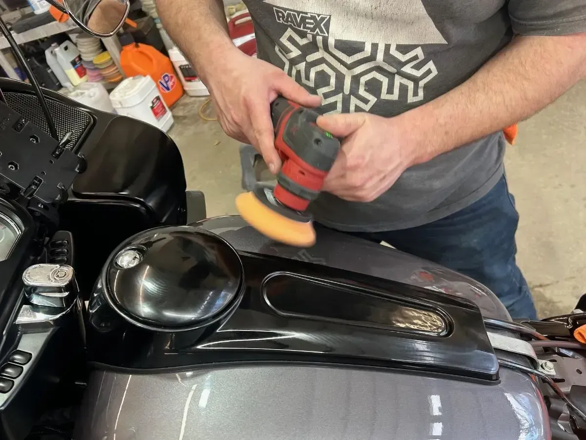 Person polishing a black motorcycle tank with a red buffer in a workshop