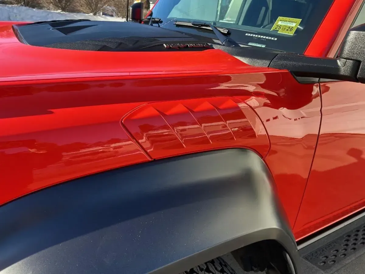 Red Ford Bronco's hood and fender with black accents. Diamond-shaped air vents on the side. Bright sunlight.