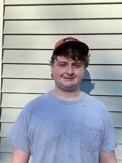 A young man wearing a hat and a grey shirt is standing in front of a house.