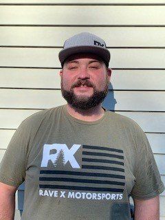 A man with a beard is wearing a rave x motorsports t-shirt and a hat.
