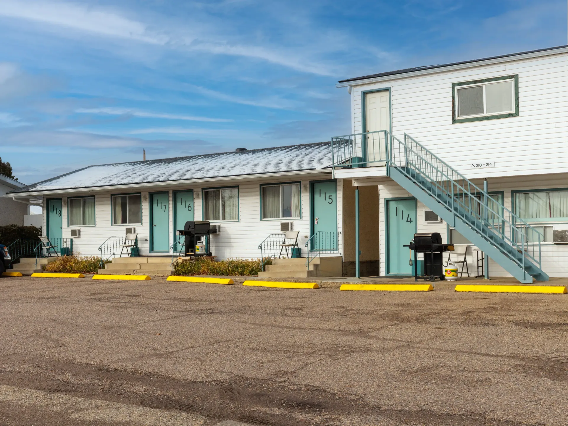 White motel building with blue doors, metal outdoor stairs, and a gravel parking lot with yellow painted curb markers.