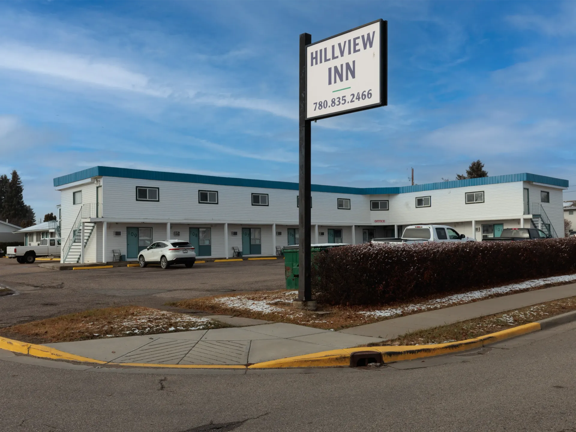 The Hillview Inn, a two-story white motel with a teal roof, located near a street corner under a blue sky.