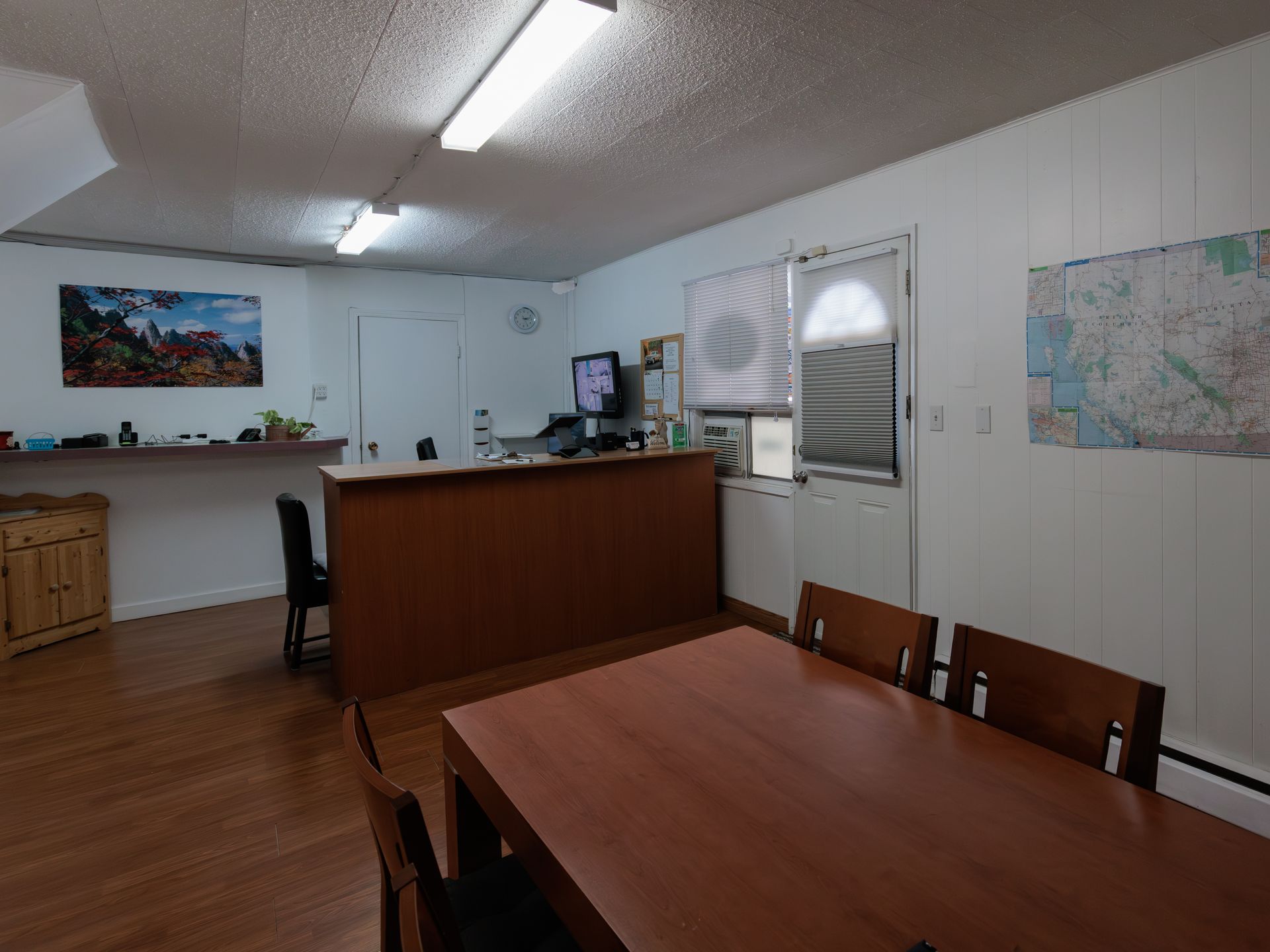 An office reception area with a wooden counter, a table with chairs, wood-look flooring, and wall-mounted maps and art.