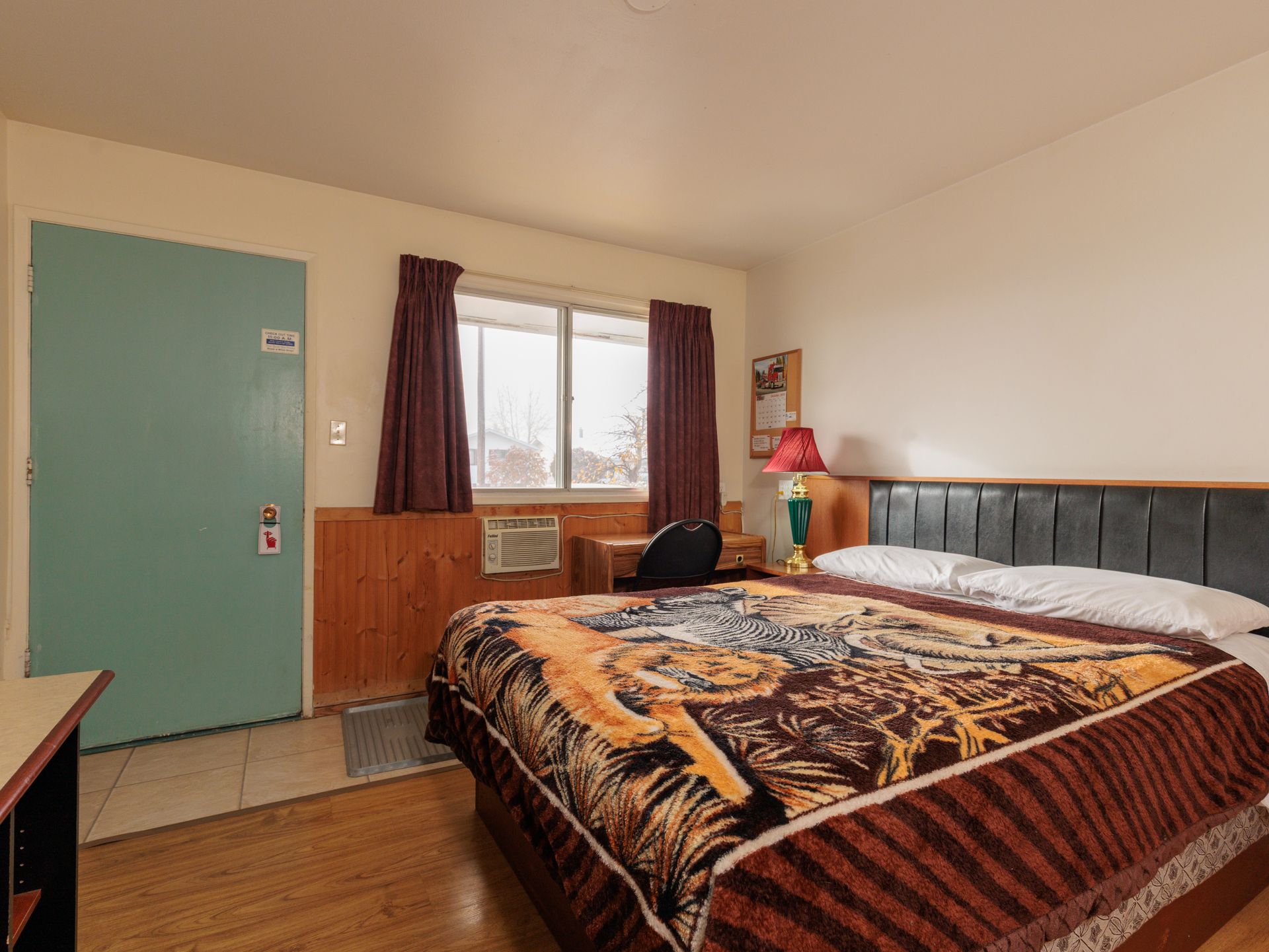 A motel room with wood paneling, a bed with a tiger-patterned blanket, a teal door, and a window with dark curtains.