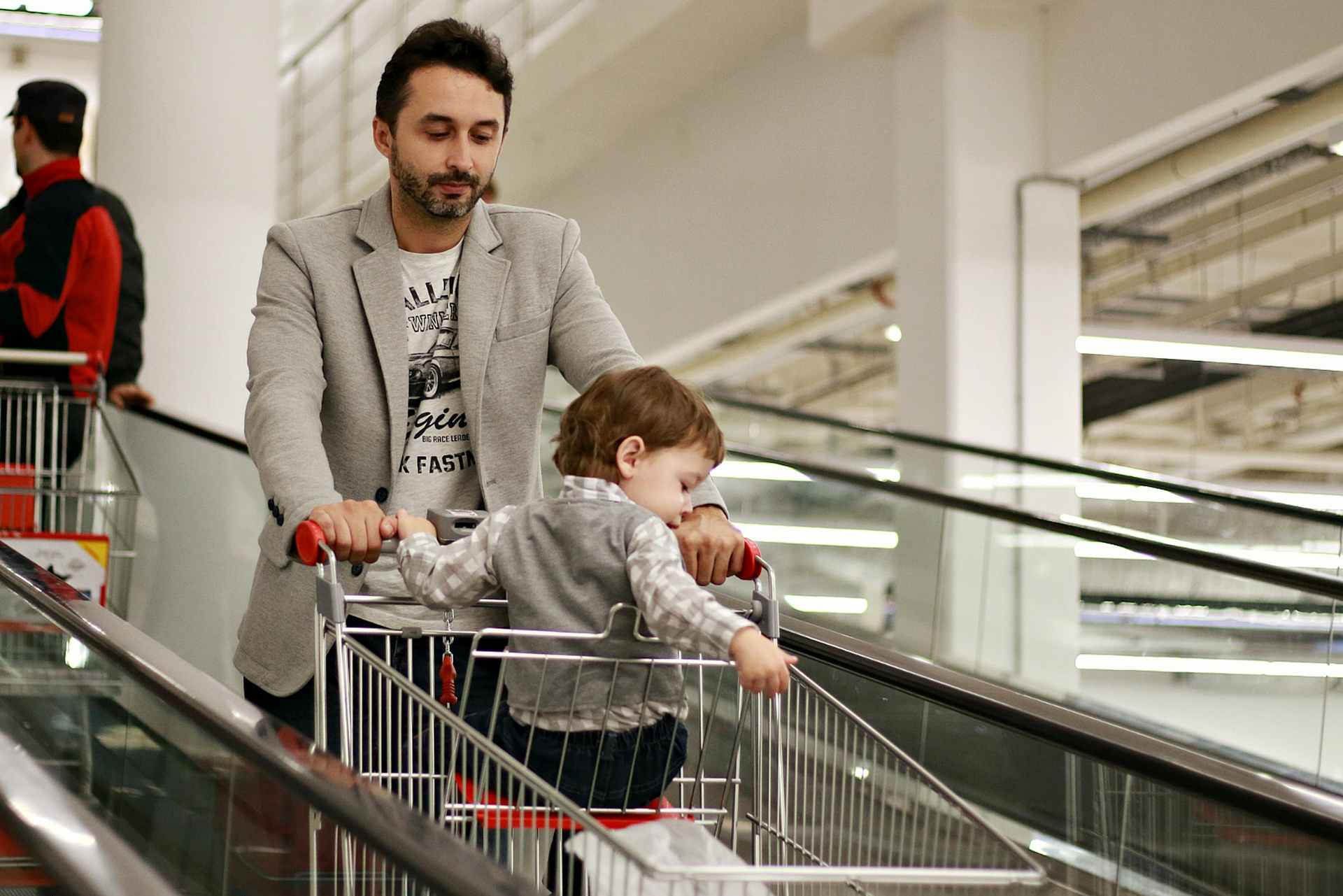 A man in a light gray blazer pushes a shopping cart with a small child seated in it while riding an escalator.