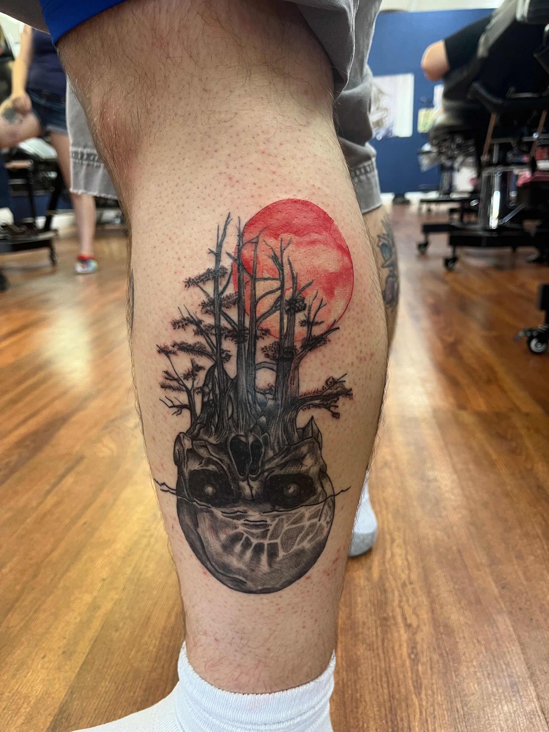 Calf tattoo of a skull with trees, a red sun, and a wooden floor background.