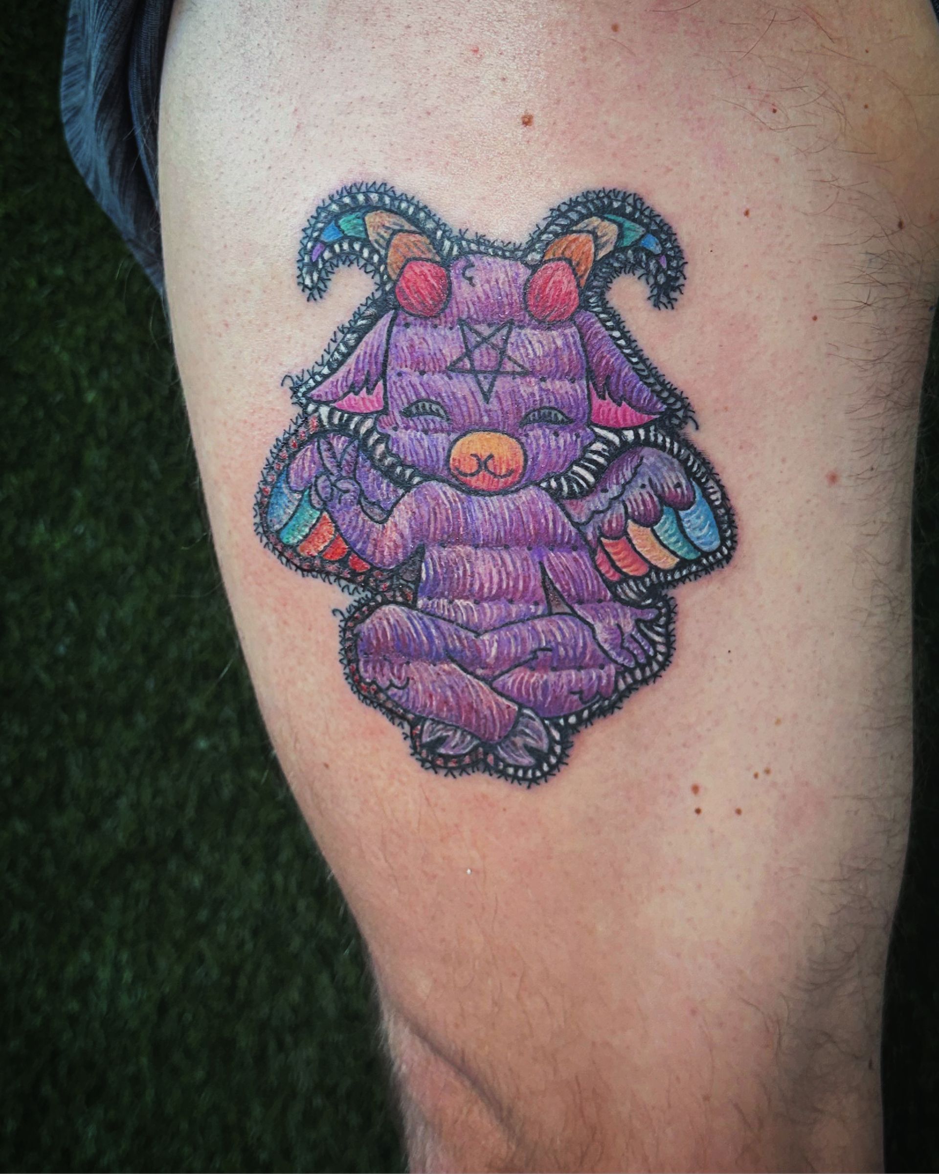 Tattoo of a purple, goat-like figure with a pentagram, sitting with folded hands, on a person’s leg.