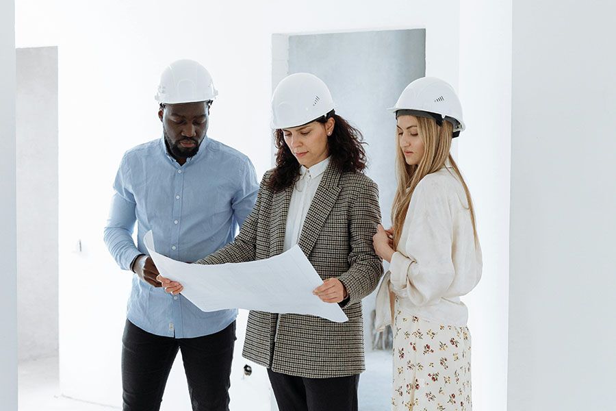 Three professionals in hard hats review a blueprint together inside a modern, unfinished white building.