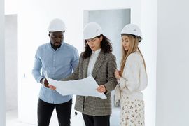 Three professionals in hard hats review a blueprint together inside a modern, unfinished white building.