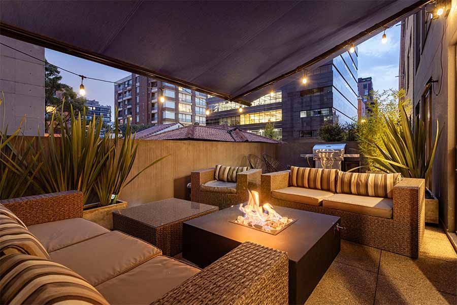 A modern rooftop terrace at dusk, featuring a fire pit table, outdoor sofas, warm string lighting, and tall plants.