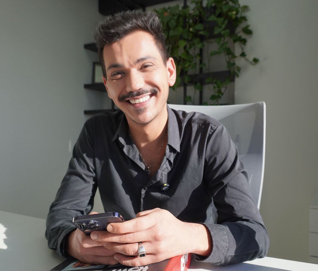 Homem com bigode, camisa escura, sorrindo, segurando o telefone em uma mesa em uma sala iluminada com uma pequena planta.