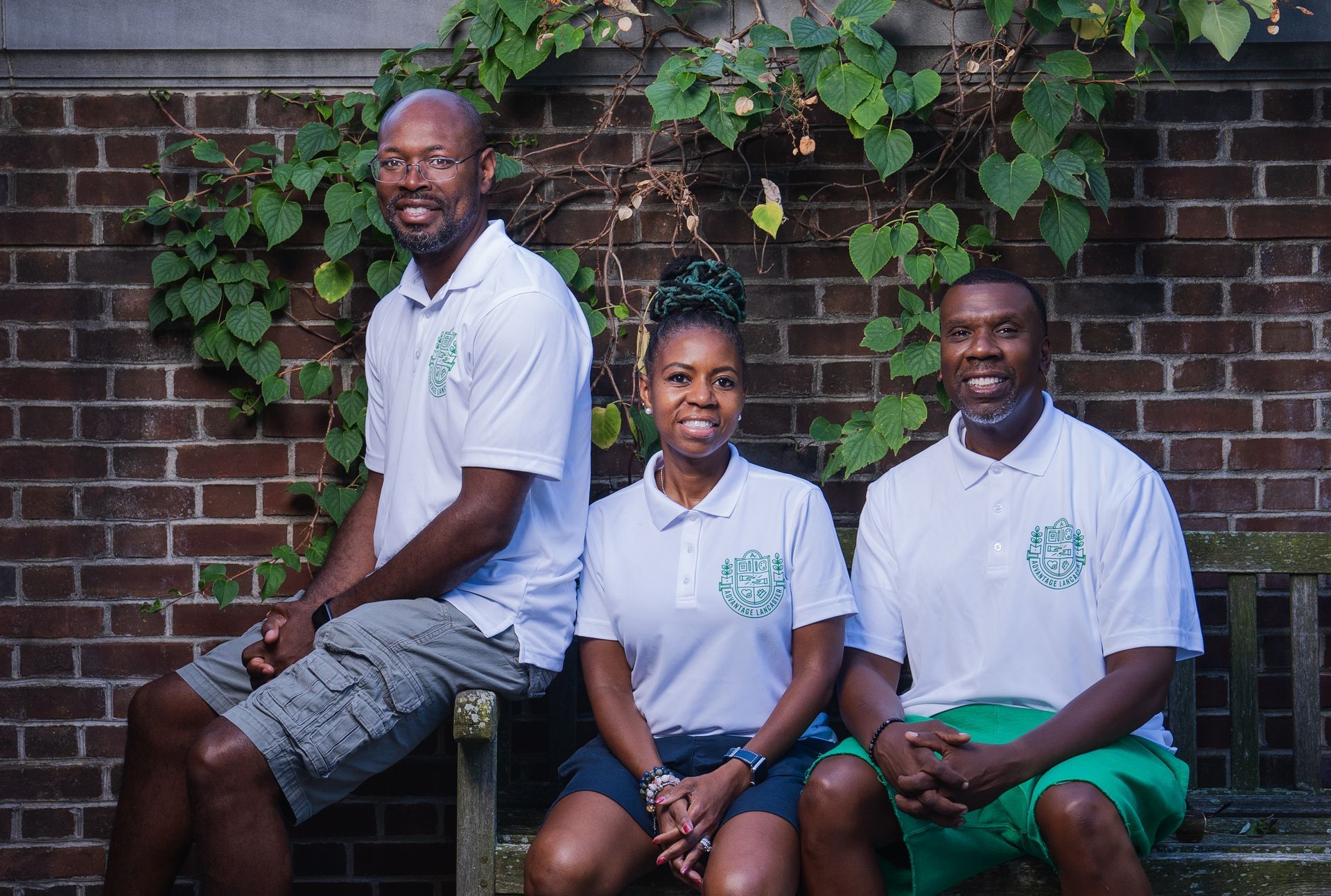 Three people are sitting on a bench in front of a brick wall.