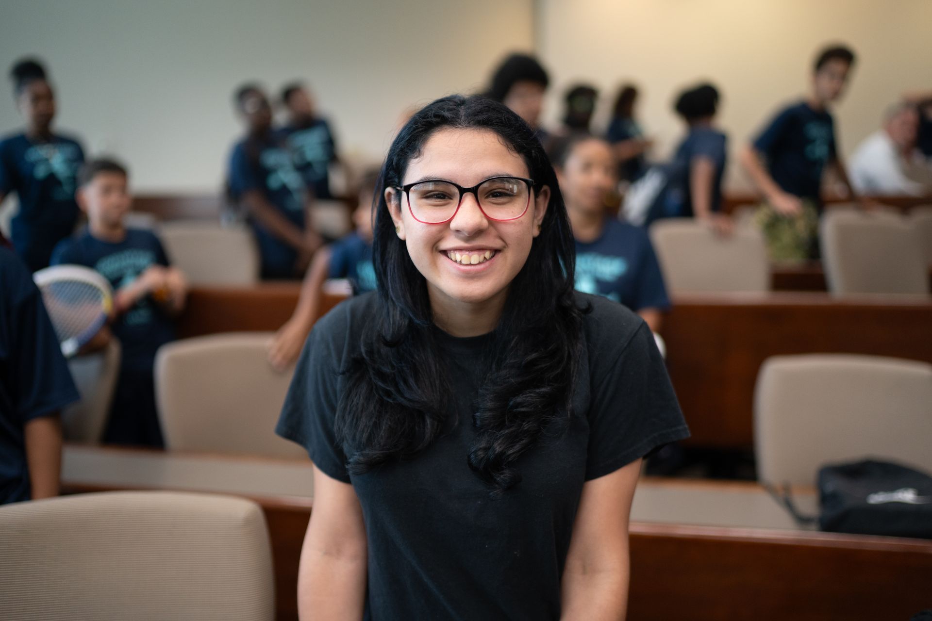 A young woman wearing glasses is smiling in a classroom.
