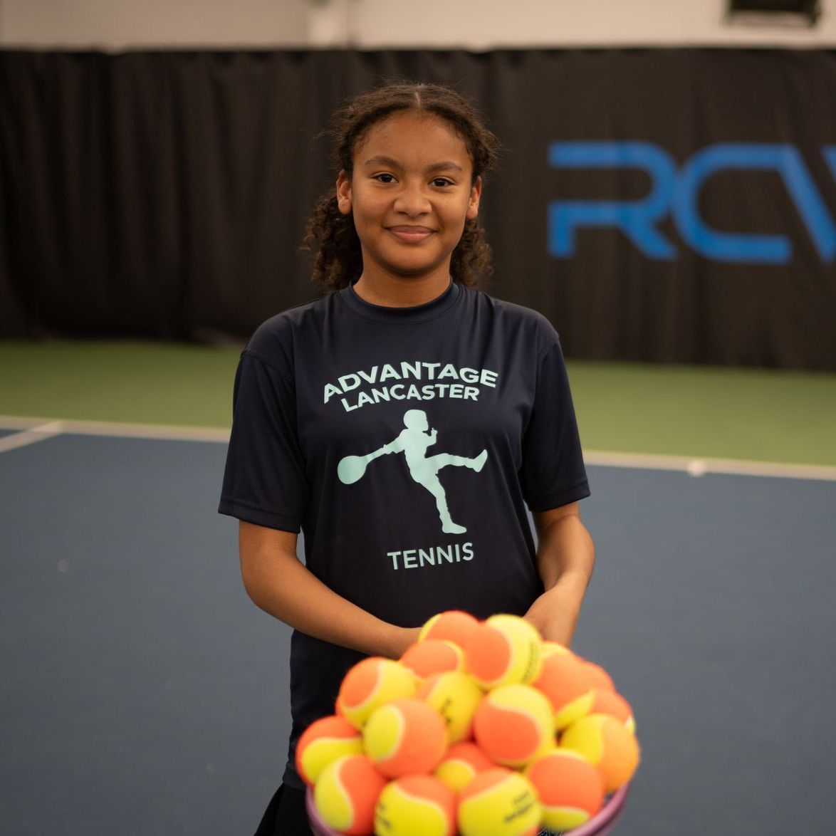 A girl wearing an advantage lancaster tennis shirt holds a bowl of tennis balls