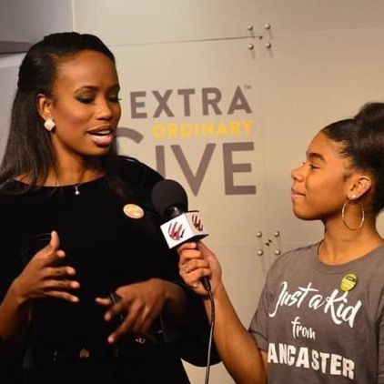 A woman is being interviewed by a girl wearing a shirt that says just a kid from lancaster