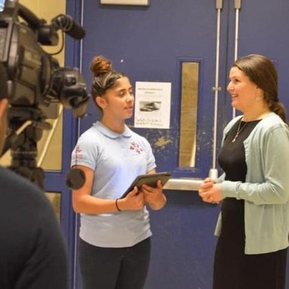 Two women are standing in front of a camera and one is holding a tablet