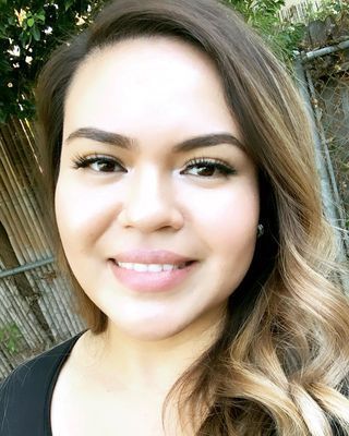 A smiling person with long, wavy, highlighted hair against an outdoor background with a fence and greenery.
