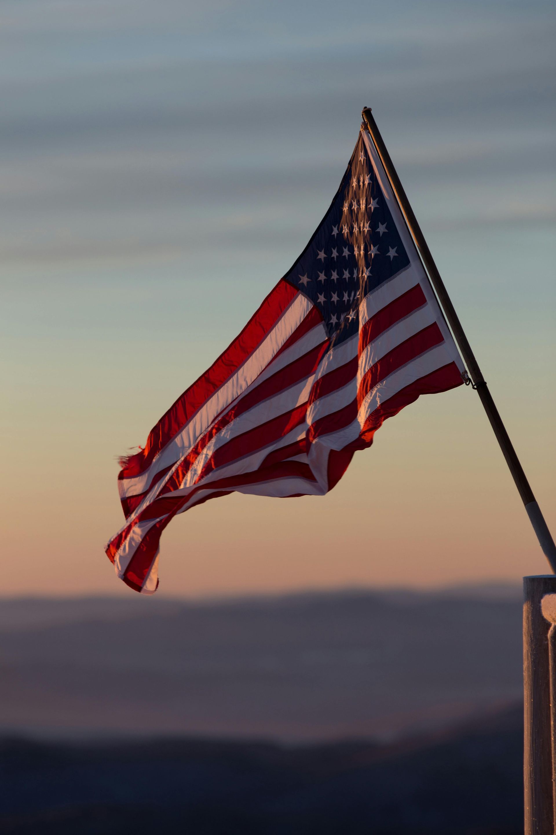 The American flag waves against a golden, sunset-colored sky above a distant, hazy mountain landscape.
