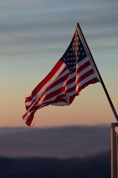 The American flag waves against a golden, sunset-colored sky above a distant, hazy mountain landscape.