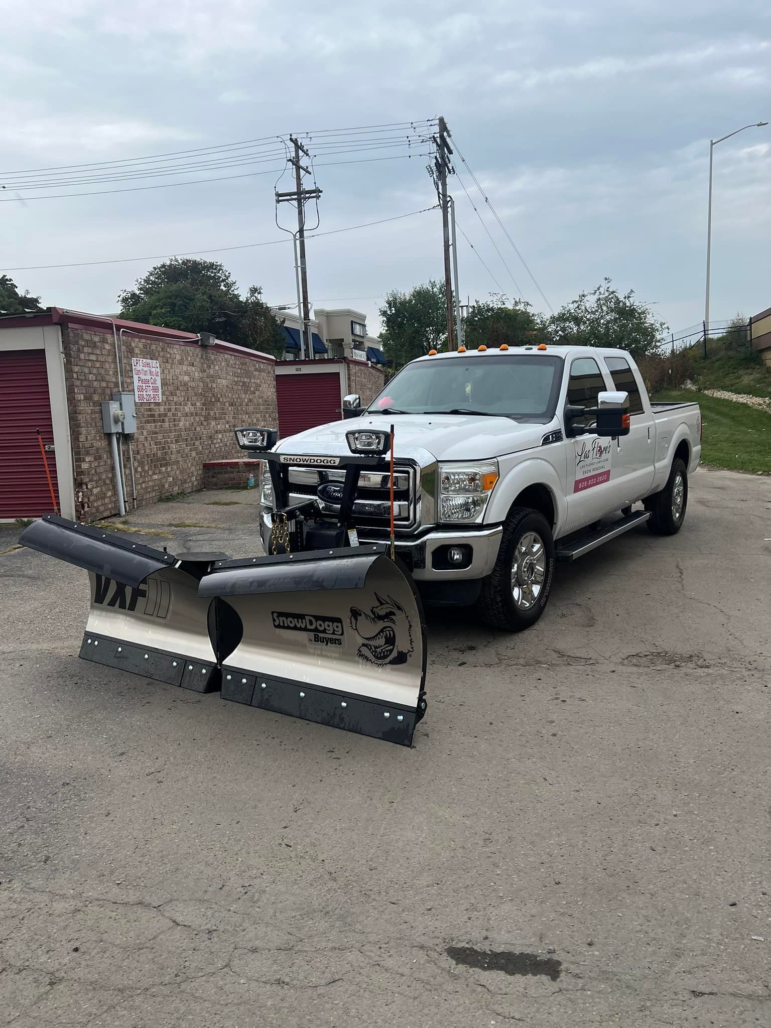 A white truck with a snow plow attached to the front is parked in a parking lot.