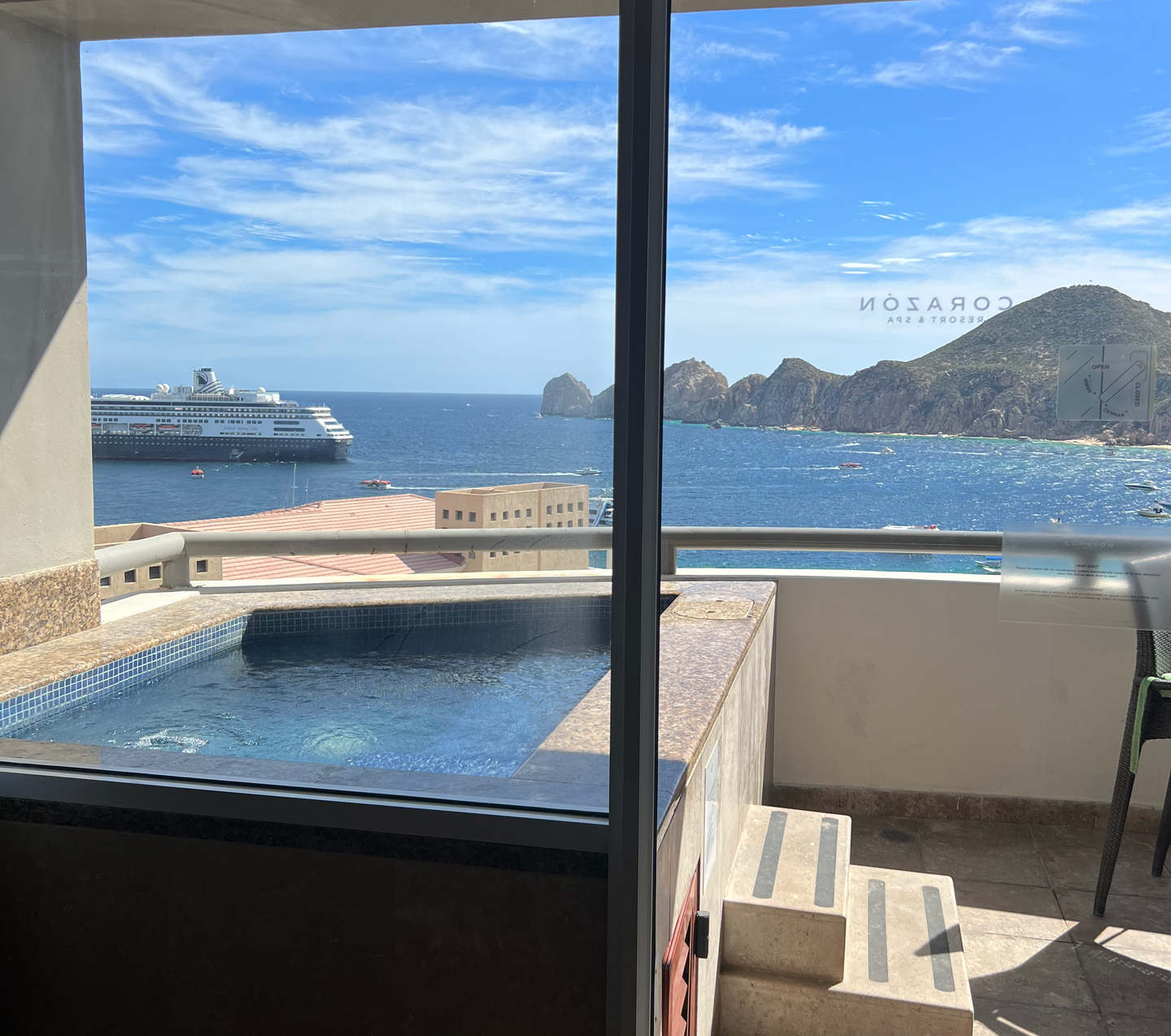 Balcony with a small pool overlooking the ocean, a cruise ship, and rocky coastline under a blue sky.