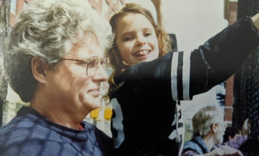 A man with white hair and wearing glasses (left) holding his daughter (right) who is smiling at the camera while pointing in the direction her father is gazing in.