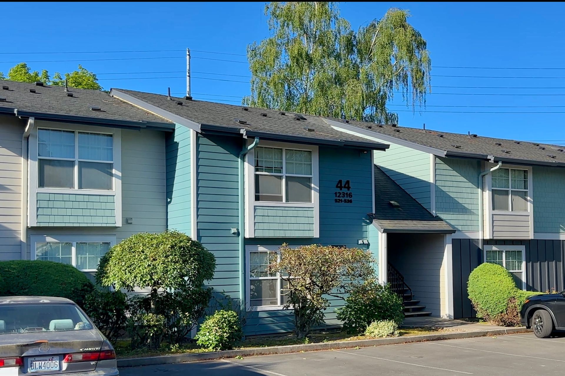 Blue two-story apartment building with parked cars and shrubs under a clear sky