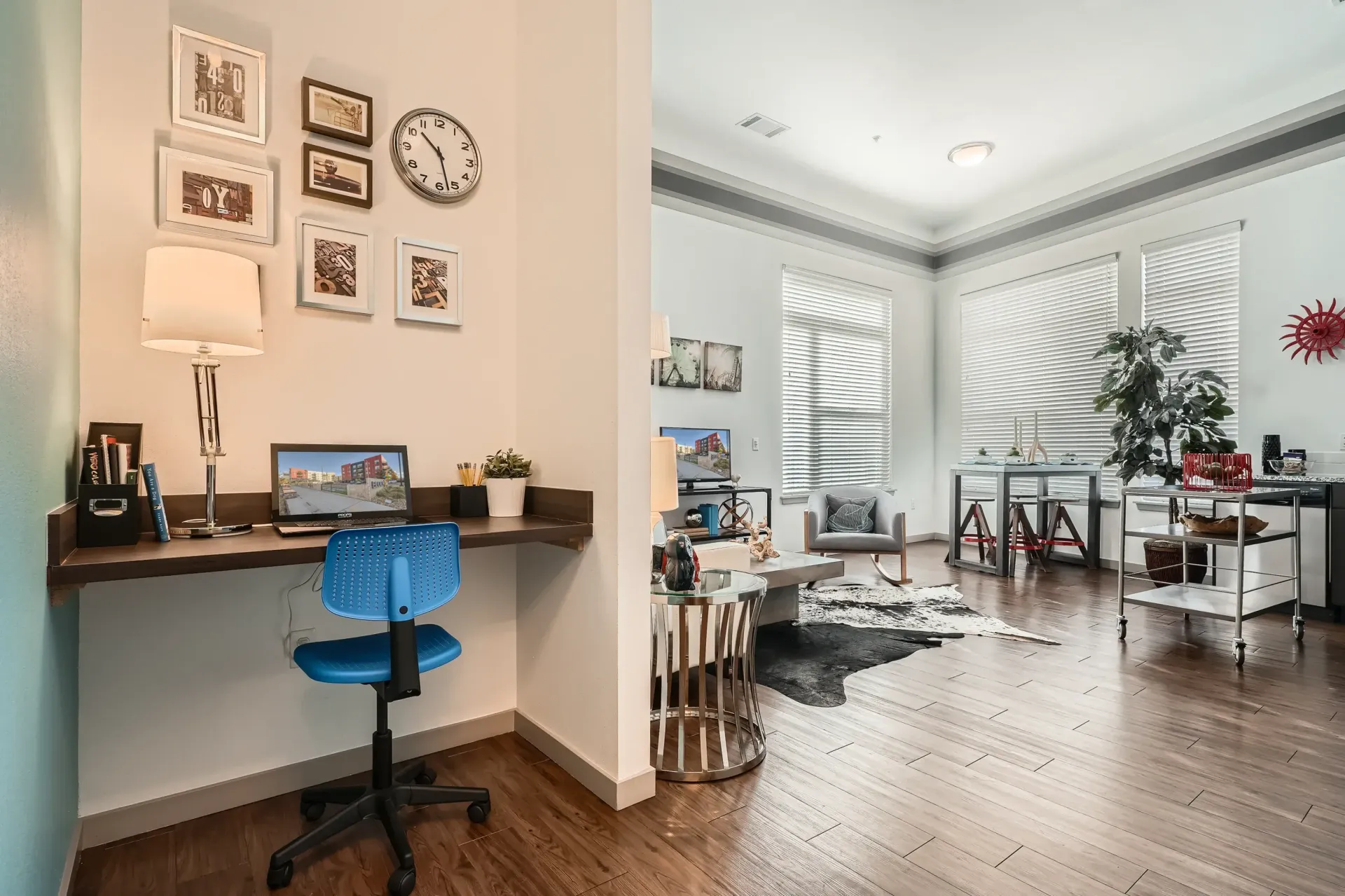 Open-plan apartment living area with a workspace nook, blue chair, sofa, coffee table, and large windows with blinds.