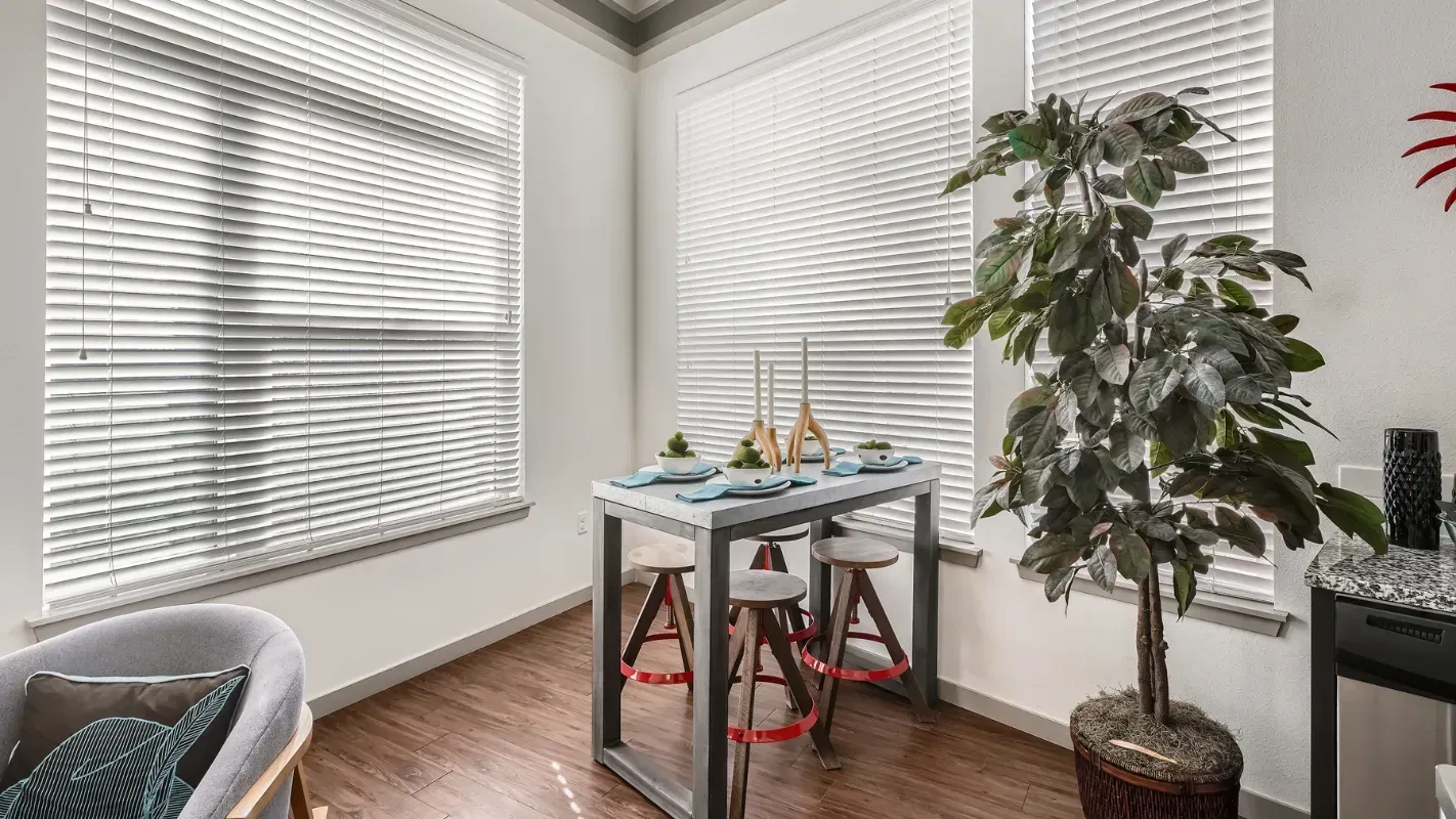 Bright apartment dining nook with a metal-frame high table, four stools, white blinds, and a tall potted plant.