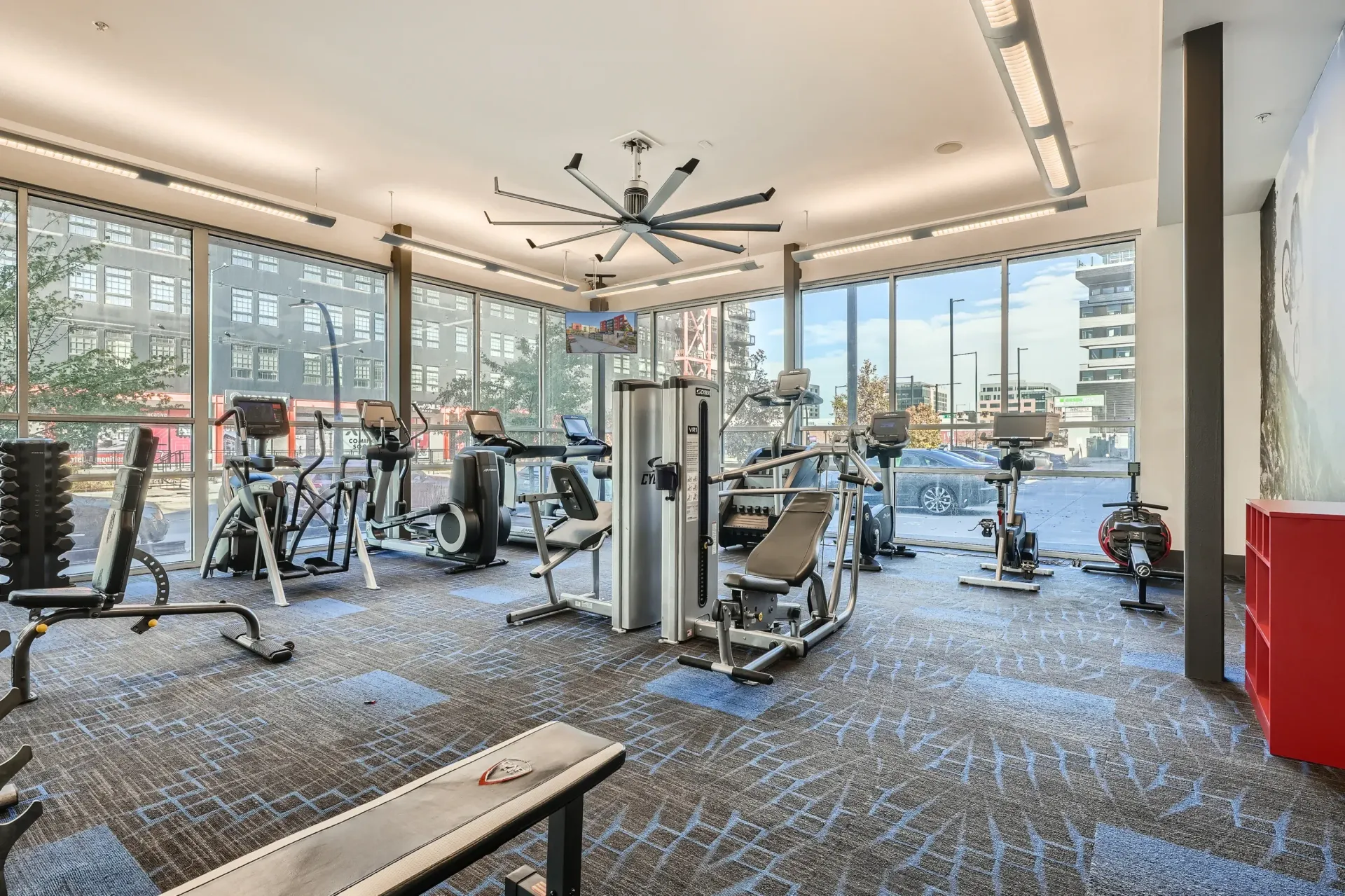 Bright communal gym with treadmills, bikes, ellipticals, and weight machines by large windows.