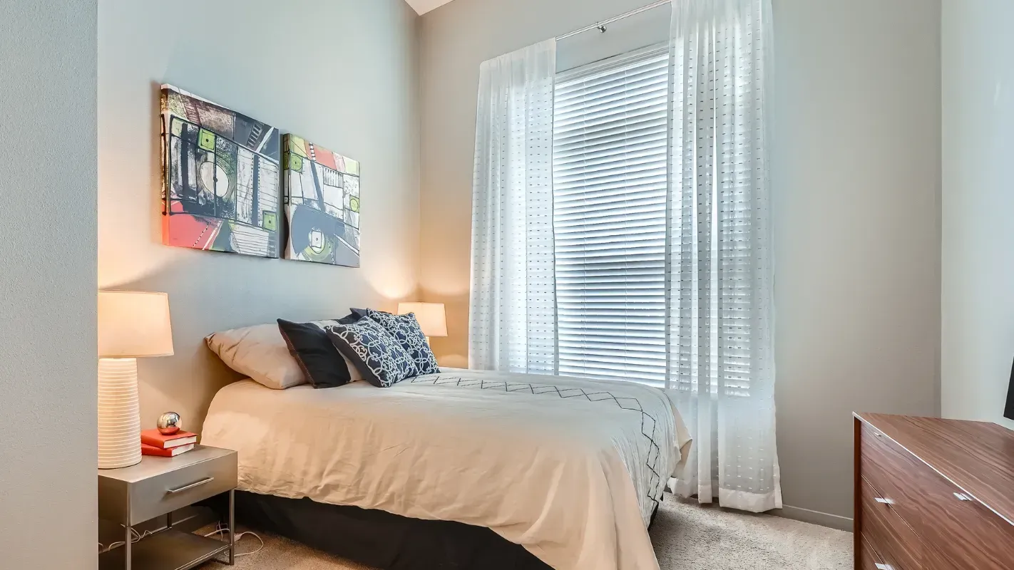 Bedroom in a modern apartment with a bed, two lamps, a nightstand, and a large window with blinds.
