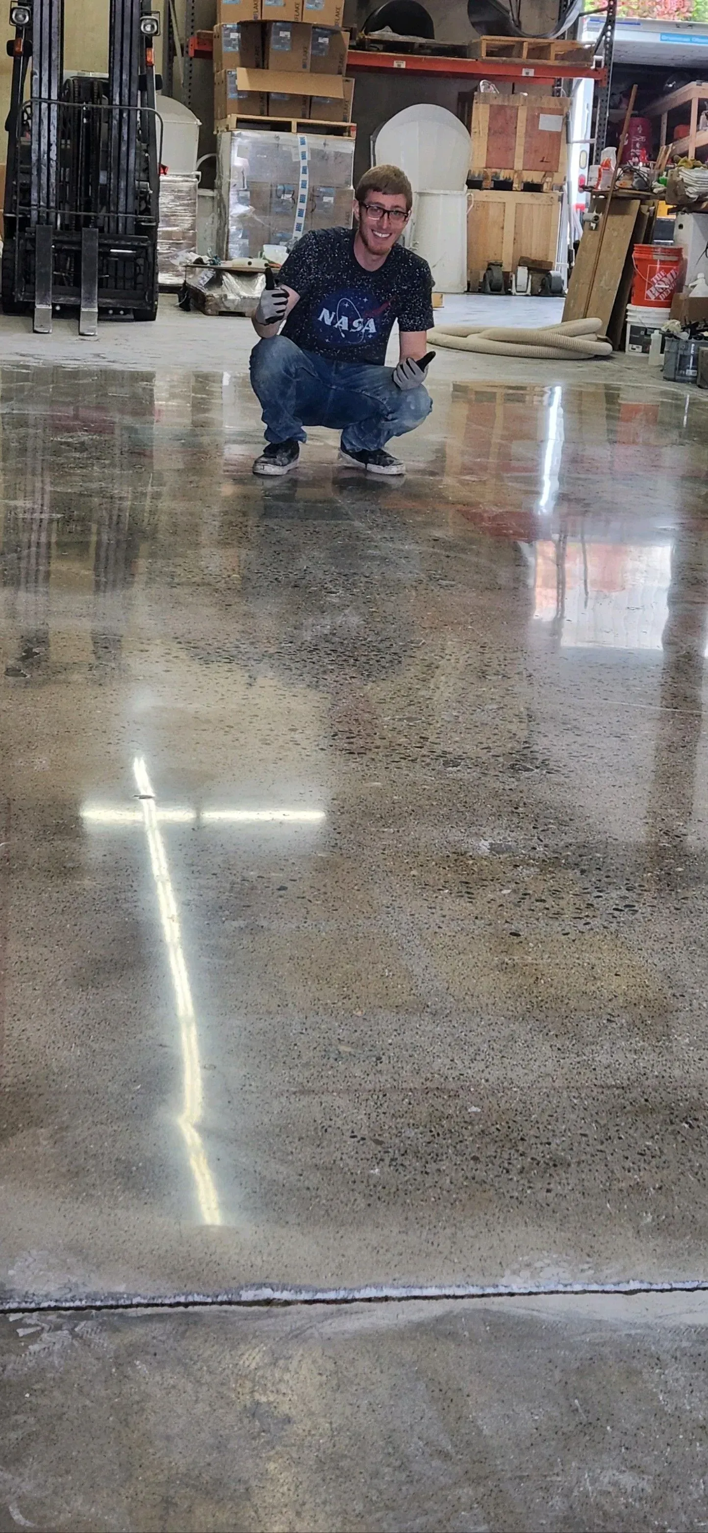 Person squatting on shiny concrete floor, likely in a warehouse. Reflections of lights on the floor.