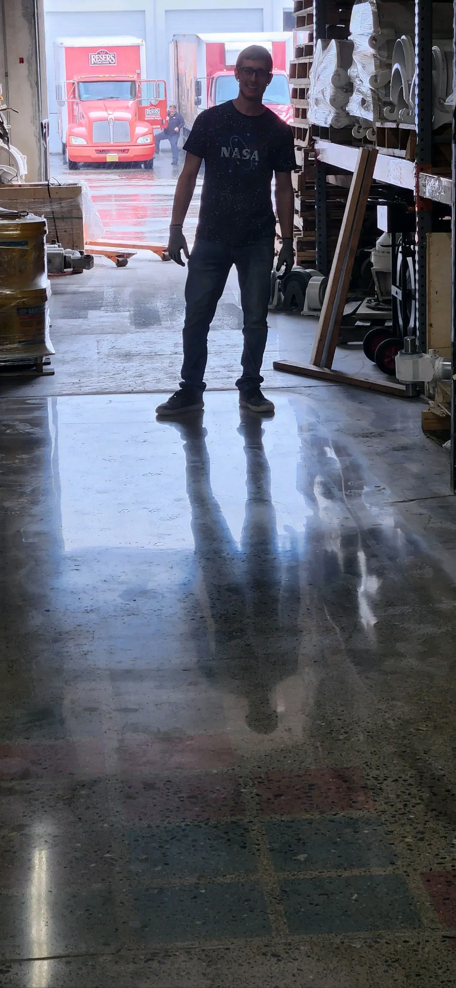 A person standing in a warehouse, reflected on the shiny concrete floor, with trucks visible in the background.