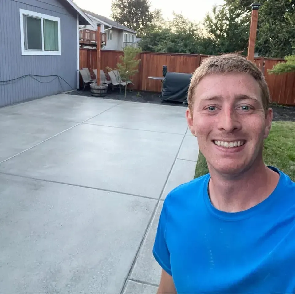 Man smiles in blue shirt, in front of a gray concrete patio, house, and wooden fence.