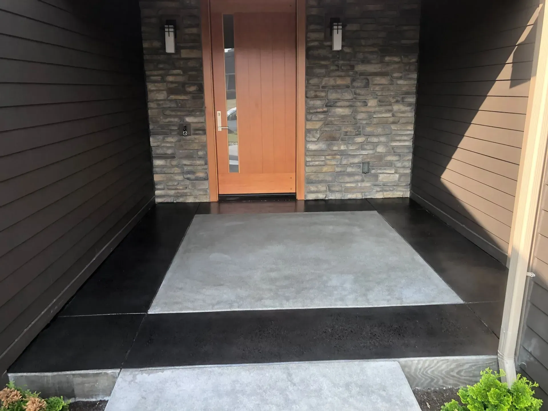 Entryway with brown door, gray concrete, and dark siding, lit by sunlight.