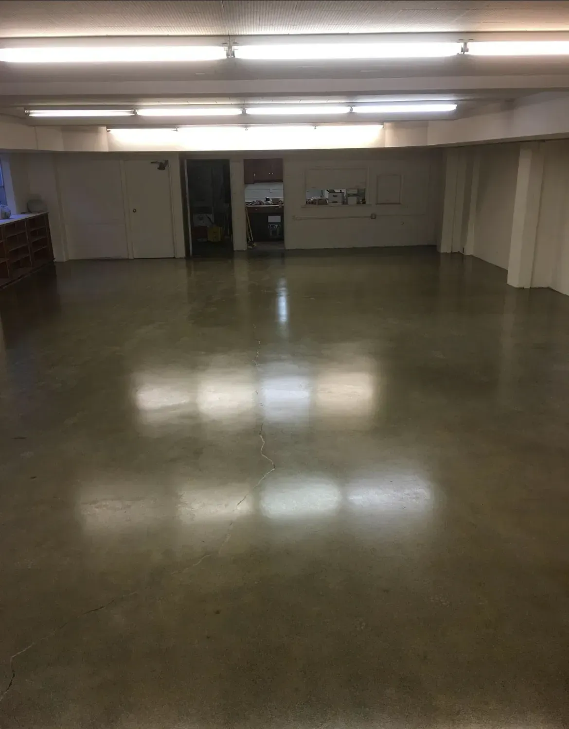 Empty room with polished concrete floor, bright overhead lighting. Beige walls, closed doors, small opening.