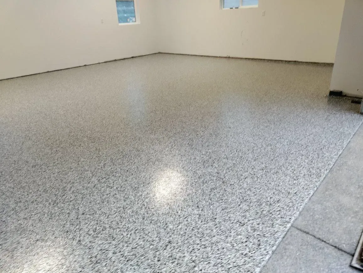 Epoxy-coated floor in a room, speckled gray and white with a glossy finish. Beige walls, windows visible.