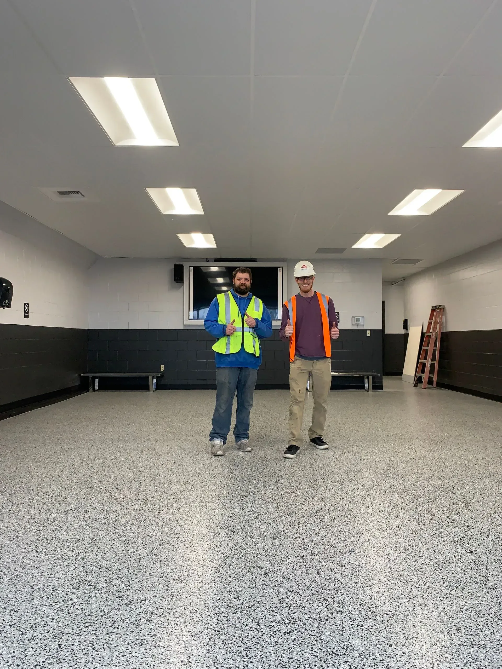 Two men in safety vests stand in a large room with a speckled floor. One holds a tablet.