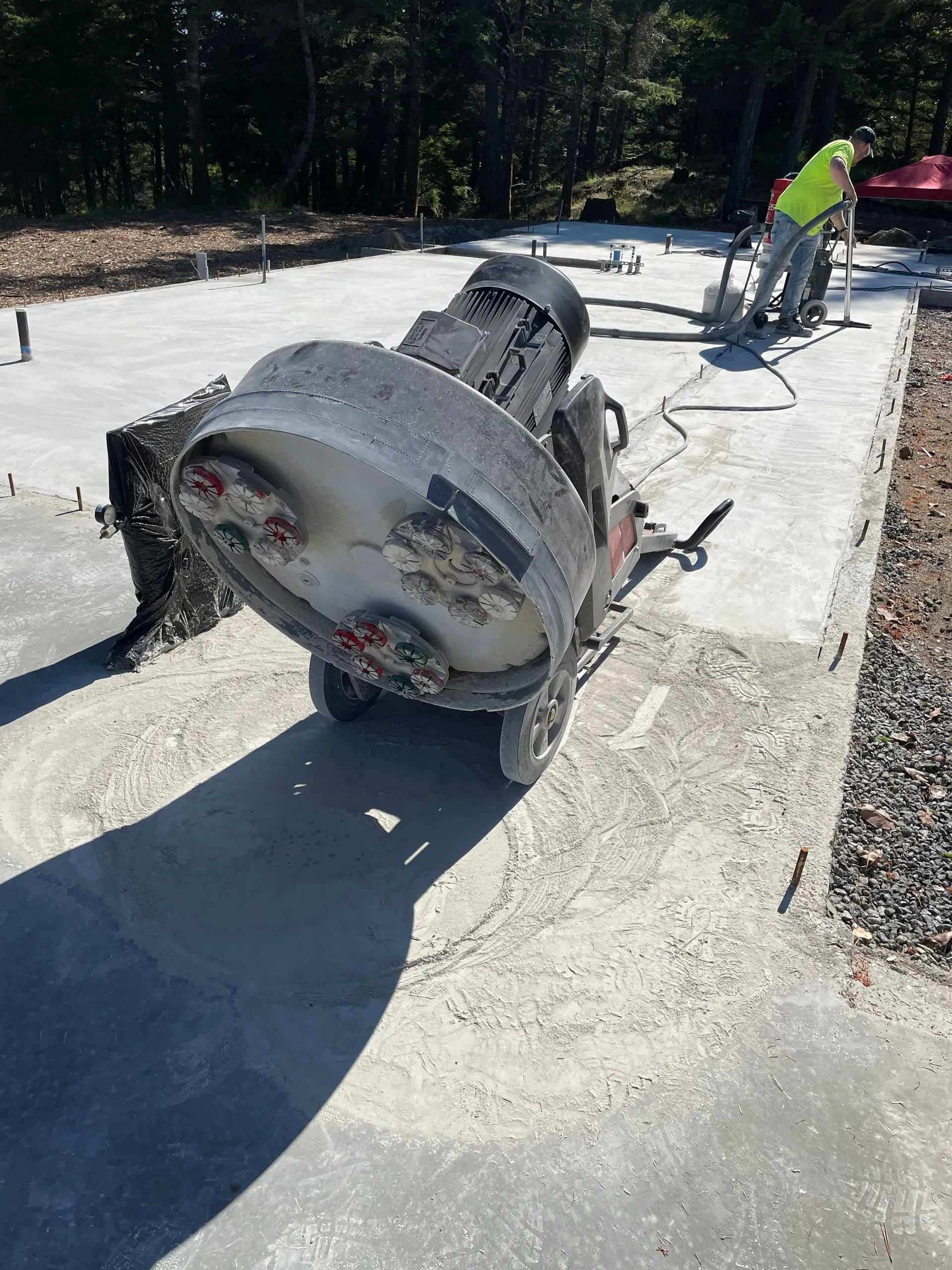 Concrete grinding machine on a concrete slab; worker in the background. Outdoors, sunny.