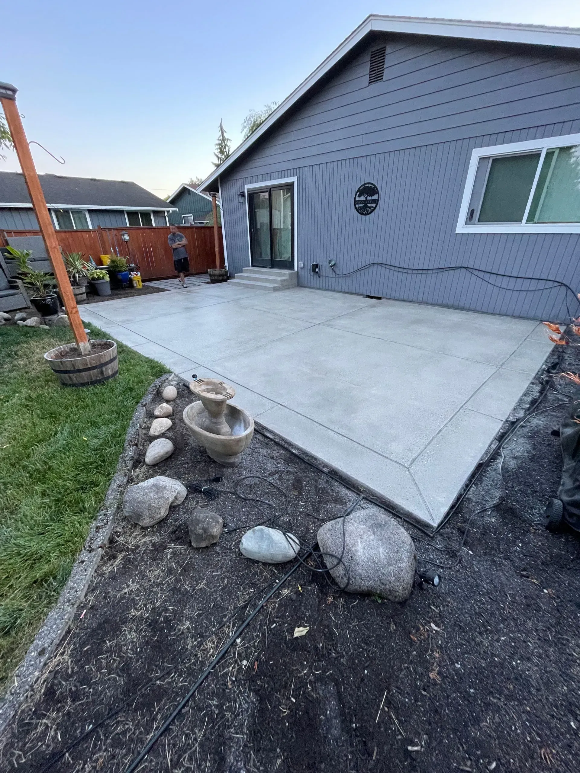 Concrete patio next to a house with a small fountain, surrounded by rocks and grass.