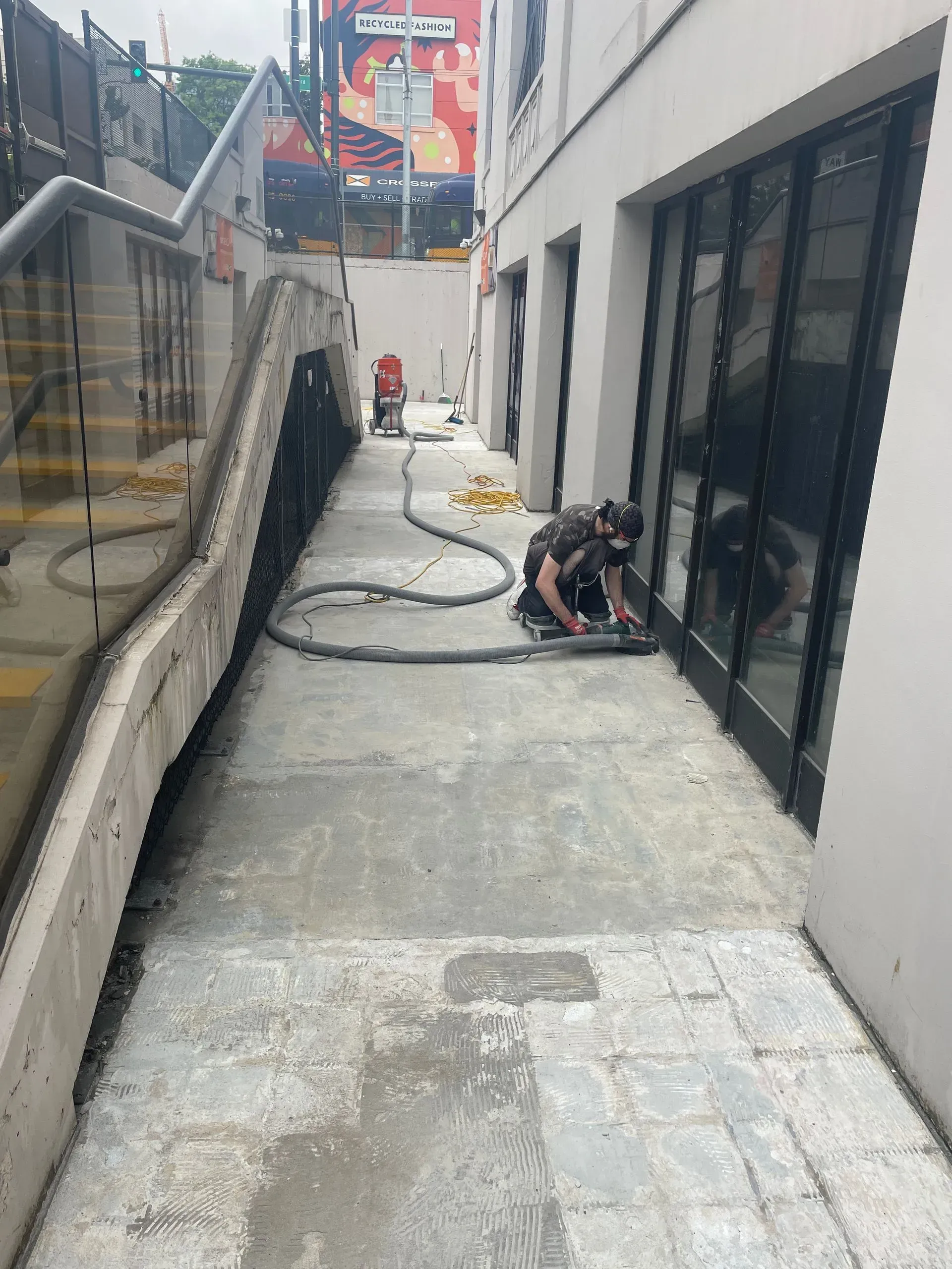 Person using a saw to cut along a glass door frame on a narrow sidewalk, near a stairwell.