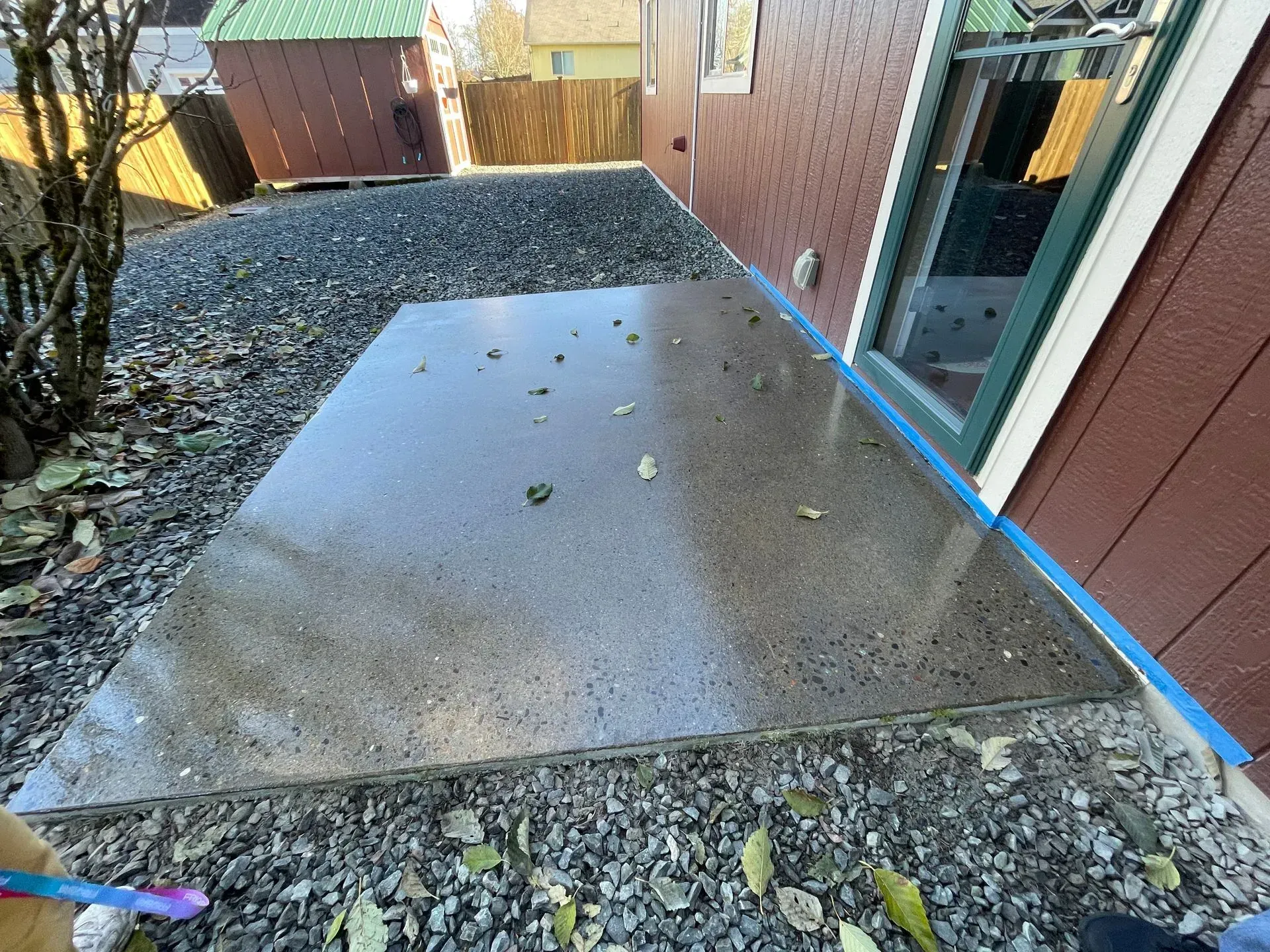 Concrete patio with gray gravel, next to a brown house with a green-framed window.