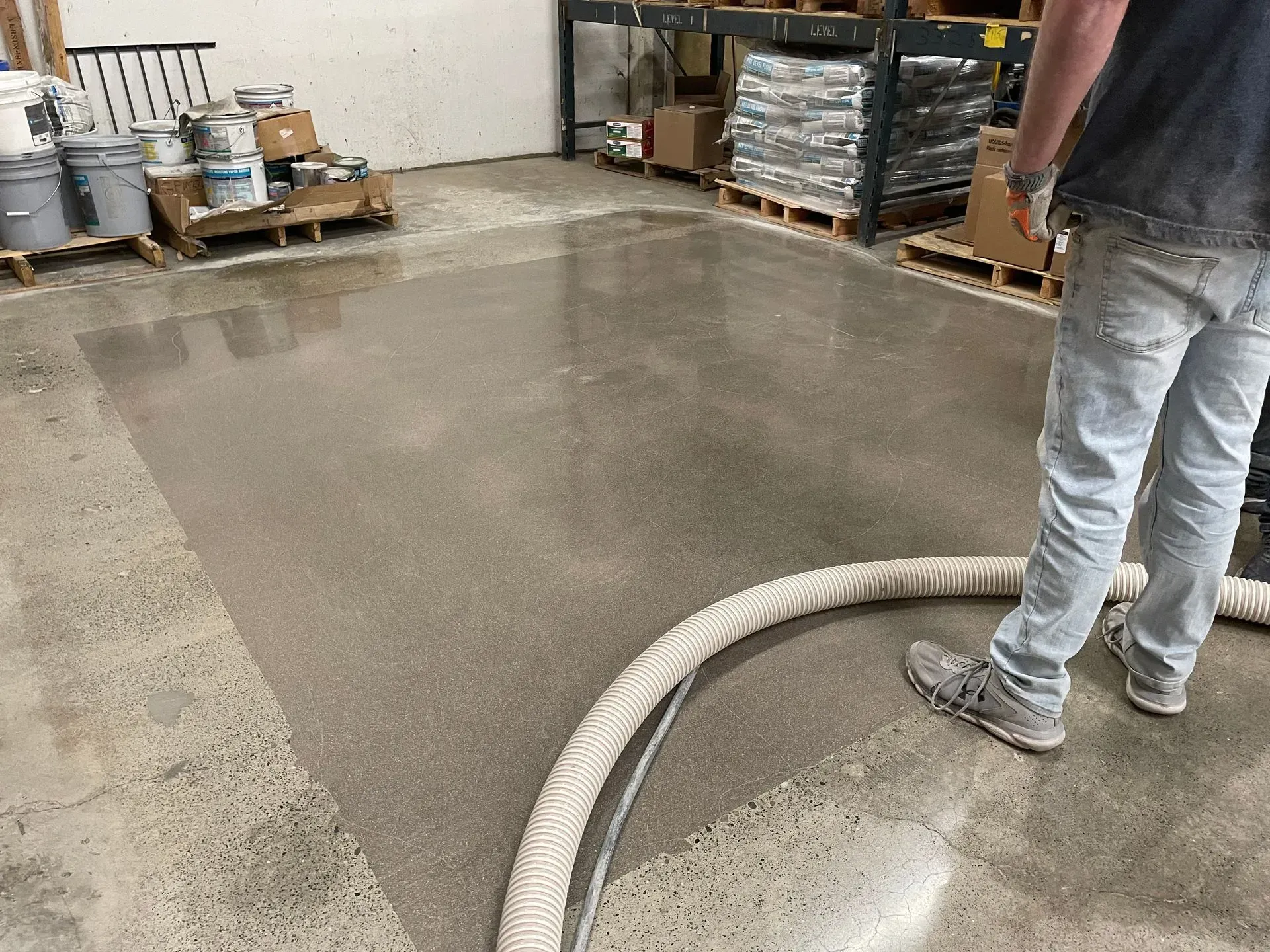 Man spraying a wet concrete floor in a warehouse. Gray flooring, coiled hose, paint cans, and pallets visible.