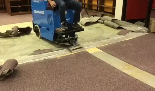 Person operating a blue carpet removal machine in a room; carpet partially removed.