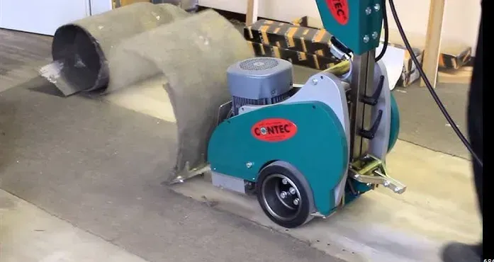 Green and gray floor grinding machine in operation, removing a coating from concrete.