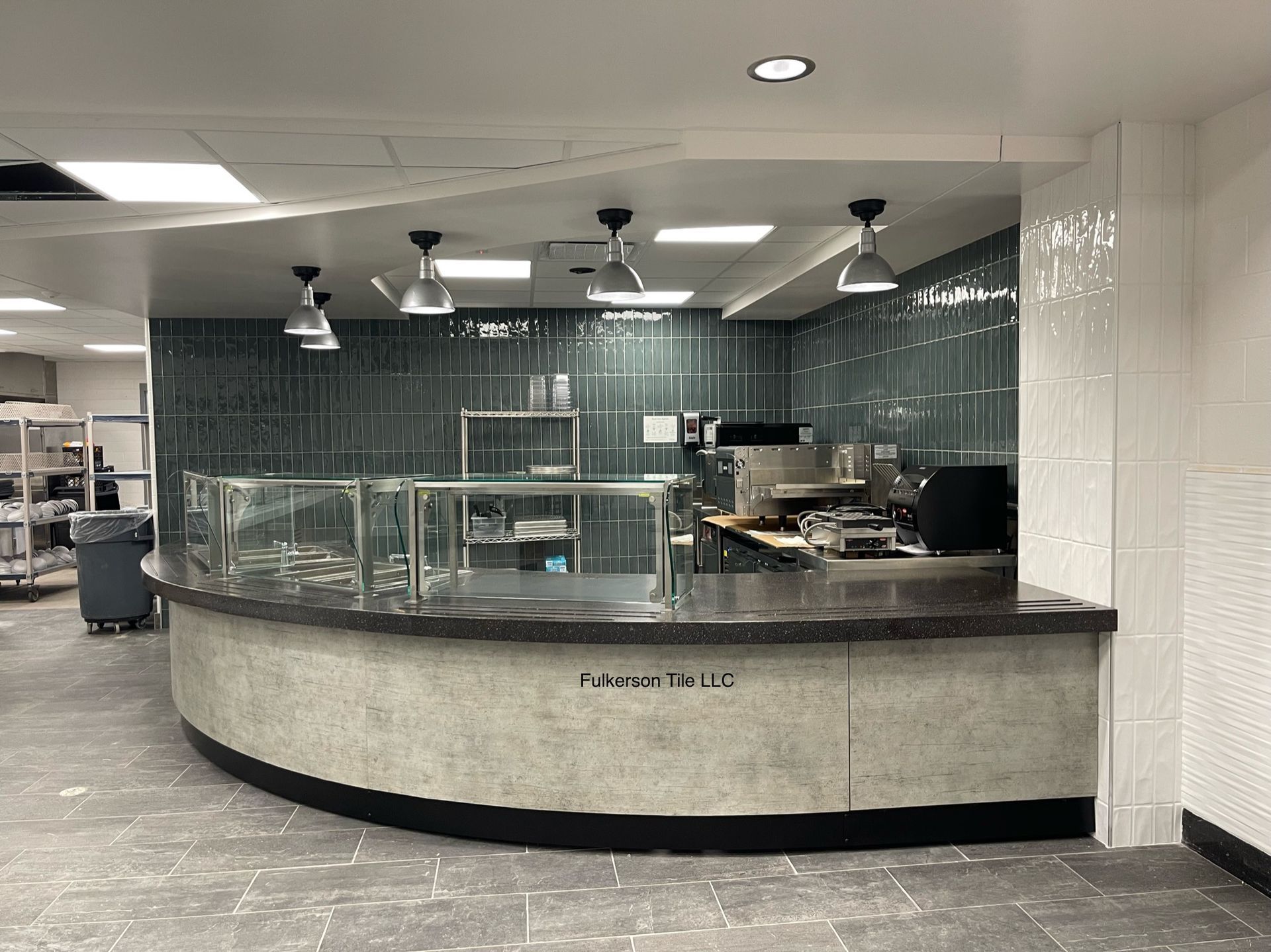 Counter with food serving area, dark countertop, aqua-green tiled wall,  stainless steel appliances, black and white floor.
