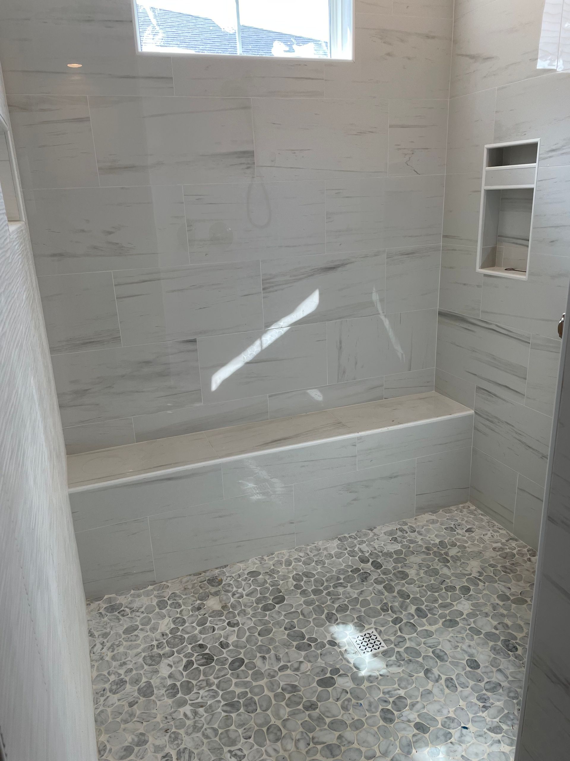 Newly tiled shower with built-in bench and recessed shelf. Grey and white stone tiles.